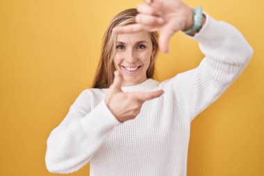 Beyaz süveter giyen, sarı arka planda gülümseyen, elleri ve parmakları mutlu yüzlü beyaz bir kadın. yaratıcılık ve fotoğrafçılık kavramı. 