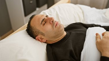Modern bir yatak odası ortamında gözleri kapalı bir yatakta dinlenen İspanyol bir adam.