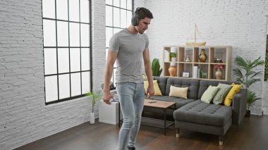 Gündelik kıyafetli genç bir İspanyol, modern, iyi dekore edilmiş bir oturma odasında dikkatlice volta atarken kulaklıkla müzik dinliyor..