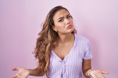 Pembe arka planda duran Latin kökenli genç bir kadın. Kolları açık ve kafası karışık, hiçbir fikri yok.. 