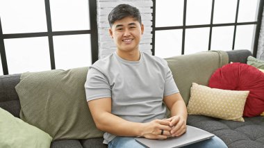 Handsome asian man smiling while seated on a couch indoors with a laptop, exuding a casual and comfortable ambiance. clipart