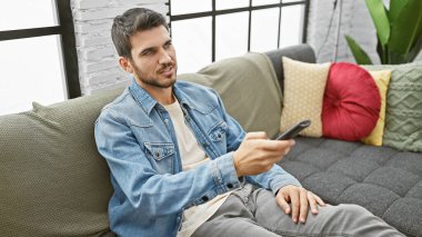 Modern oturma odasında uzaktan kumandayla kanepeye uzanan günlük giysili yakışıklı İspanyol bir adam.