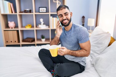 Genç İspanyol adam akıllı telefonda konuşuyor. Yatak odasında kahve içiyor.