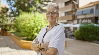 Kendinden emin, gri saçlı, gülümseyen, güneşli parkta duran, kollarını kavuşturan, eğlenceli, olgun bir yaşam tarzı yaşayan, neşeli, doğanın yeşilliğinin tadını çıkaran son sınıf kadını.