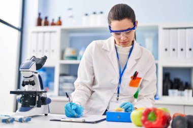 Young beautiful hispanic woman scientist weighing piece of apple writing report at laboratory
