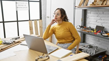 Sarı kazaklı düşünceli bir kadın marangozluk atölyesinde dizüstü bilgisayar ve aletlerle proje analiz ediyor..