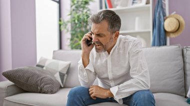 Stresli, gri saçlı, evinde kanepede oturan, hararetli bir anlaşmazlıkla telefonda ciddi şekilde tartışan İspanyol asıllı genç bir adam.