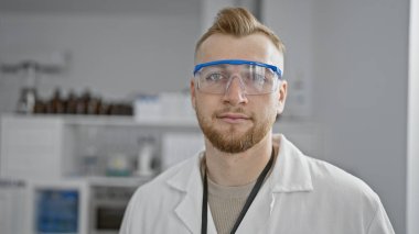 Sakallı, gözlüklü, laboratuvar ortamında laboratuvar önlüğü giyen, profesyonel ve odaklanmış görünen beyaz bir adam..