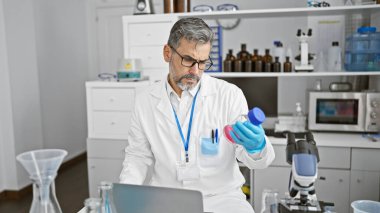 Laboratuvarda laptopla test tüpü tutan beyaz saçlı genç bir bilim adamı.