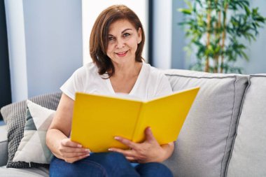 Orta yaşlı kadın kitap okuyor. Evdeki kanepede oturuyor.