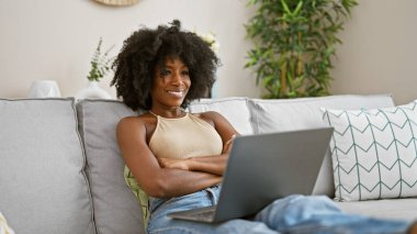 Kanepede oturan, dizüstü bilgisayar kullanan Afro-Amerikan kadın evinde kollarını kavuşturup el kol hareketi yapıyor.