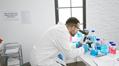 Laboratuvar ortamında laboratuvar önlüğü ve eldivenleriyle mikroskopla örnekleri analiz eden İspanyol bilim adamı..