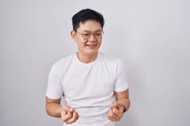 Beyaz arka planda duran Asyalı genç adam barış sembolü yapıyor. Yüzünde parmaklar, gülümseyerek zaferi gösteriyor. 