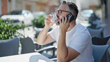 Gri saçlı, kahve içen, kahve dükkanının terasında akıllı telefondan konuşan adam.