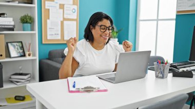 Neşeli İspanyol bir kadın modern ofis masasında başarısını dizüstü bilgisayar ve malzemelerle kutluyor..