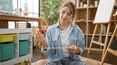 Genç kadın sanatçı resim stüdyosunda yere oturmuş palete renk döküyor.