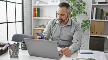 Modern bir ofiste akıllı telefon ve dizüstü bilgisayar kullanan sakallı, olgun bir İspanyol..