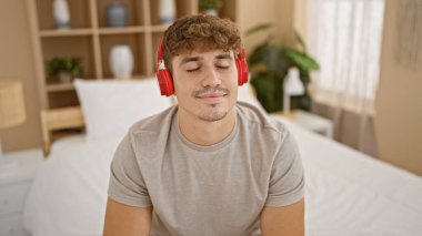 Genç, yakışıklı İspanyol adam rahat bir şekilde rahatlıyor, neşe içinde eğlenceli müzikler dinliyor, sade yatak odasında rahat bir yatakta oturuyor, mutluluğu ve güveni somutlaştırıyor ve sabahın tadını çıkarıyor.