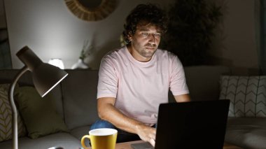 Geceleri rahat bir oturma odasında dizüstü bilgisayar kullanan odaklanmış genç bir adam, dekoratif bir lambası ve yastığı var..