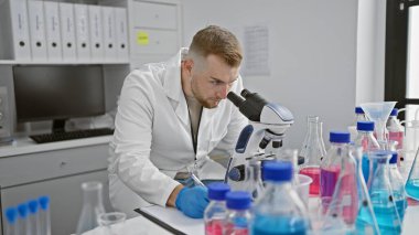 Sakallı, genç, beyaz bir adam laboratuarda çalışıyor. Mikroskopla örnekleri inceliyor..