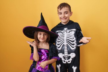 Heyecanlı cadılar bayramı kostümleri içinde neşeli gülümsemeler ve muzaffer ifadelerle zaferi kutlayan sarı bir arka plan duvarında tek başına duran küçük kardeşler.