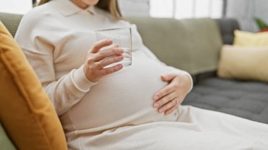 Genç bir İspanyol hamile kadın, bir kanepede dinlenirken elinde bir bardak su tutuyor. Sağlık ve anneliği simgeliyor..