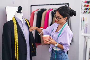 Young beautiful hispanic woman tailor drinking coffee measuring jacket at tailor shop