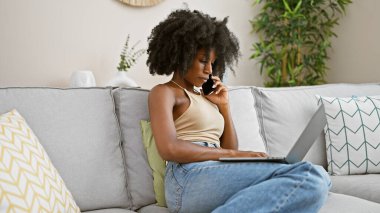 African american woman talking on smartphone using laptop at home