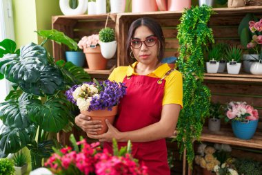 Çiçekçi dükkanında çalışan genç bir Arap kadın elinde bitki tutarken düşünceli ve ayık bir ifade takınıyor. Kendinden emin görünüyor. 