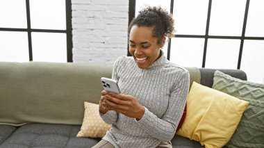 Oturma odasındaki kanepede akıllı telefon kullanan gülümseyen kadın.