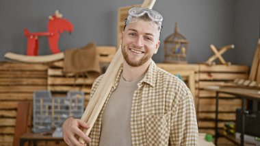 Marangozluk atölyesinde elinde tahta olan sakallı, gülümseyen genç adam.