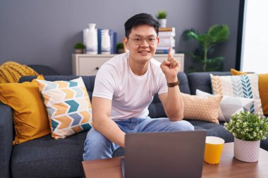 Genç Asyalı adam evinde dizüstü bilgisayar kullanıyor kanepeye oturmuş başarılı bir fikirle işaret ediyor. Heyecanlı ve mutlu. Bir numara.. 