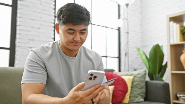 Asyalı adam akıllı telefon kullanıyor ve kredi kartını içeride tutuyor, modern oturma odasında online alışveriş veya mobil bankacılık portresi yapıyor..