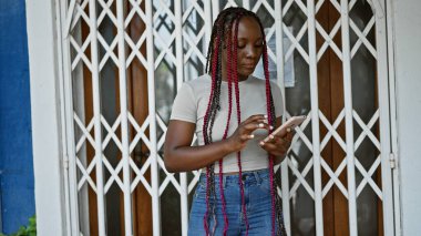 Güzel örgüler! Çarpıcı siyahi Afrikalı Amerikalı kadın ciddi ciddi akıllı telefonuna odaklanıyor, canlı güneşli şehir sokak yaşam tarzında duruyor..