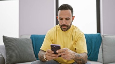 Odaklanmış, sarı gömlekli, sakallı bir İspanyol akıllı telefonunu evde, yastıklı, gri bir koltukta kullanıyor..