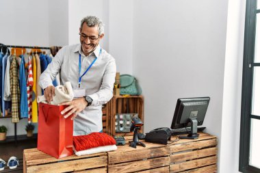Orta yaşlı, gri saçlı bir dükkan asistanı giyim mağazasından alışveriş yapmaya hazırlanıyor.