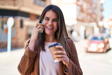 Young beautiful hispanic woman talking on smartphone drinking coffee at street