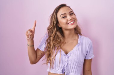 Young hispanic woman standing over pink background showing and pointing up with finger number one while smiling confident and happy. 