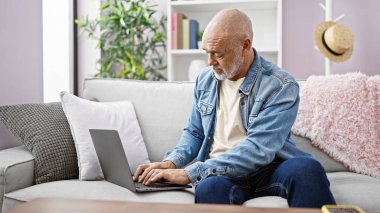 Kanepede dizüstü bilgisayar kullanan sakallı, gri saçlı, olgun bir İspanyol.