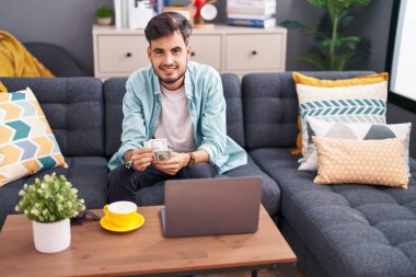 Young hispanic man using laptop counting dollars at home