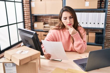 Young beautiful hispanic woman ecommerce business worker using laptop reading paperwork at office