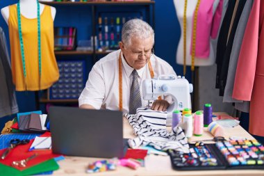 Terzide dikiş makinesi ve dizüstü bilgisayar kullanan orta yaşlı gri saçlı bir adam.