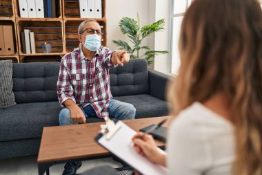 Tıbbi maske takan kıdemli bir adam kamerayı işaret ederek oturuyor ve sen kendine güvenen bir hareket yapıyorsun. Psikoloji kliniğine ciddi bakıyorsun.