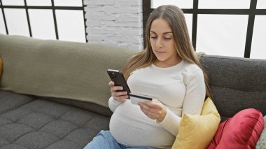 Oturma odasında kredi kartı ve akıllı telefon kullanan hamile İspanyol kadın..
