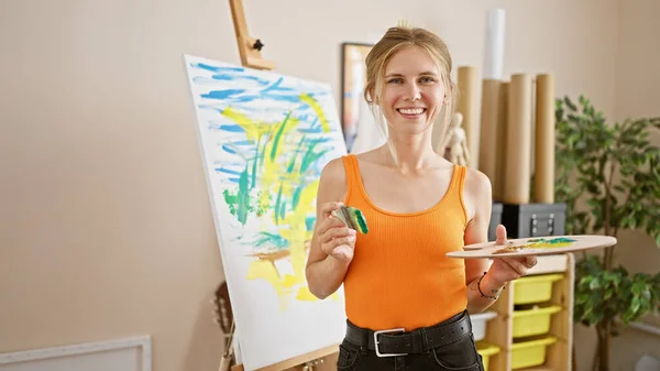 A smiling young woman artist in a studio holding a paintbrush and palette with a colorful abstract painting on the easel.