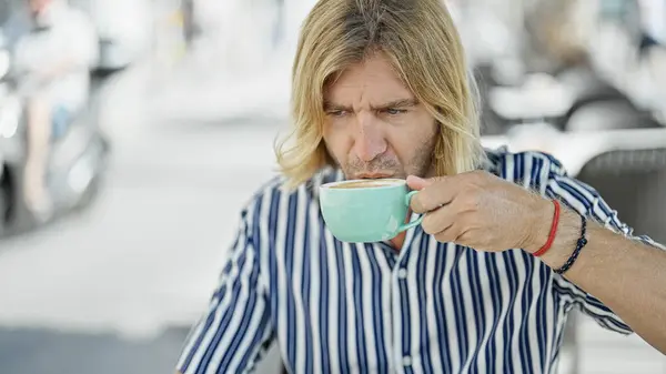 A man with long blond hair sipping coffee at an outdoor urban cafe captures a relaxed lifestyle moment.