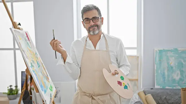 Confident young hispanic man, an artist with grey hair, joyfully stands smiling in an art studio, embracing the world of creativity and painting