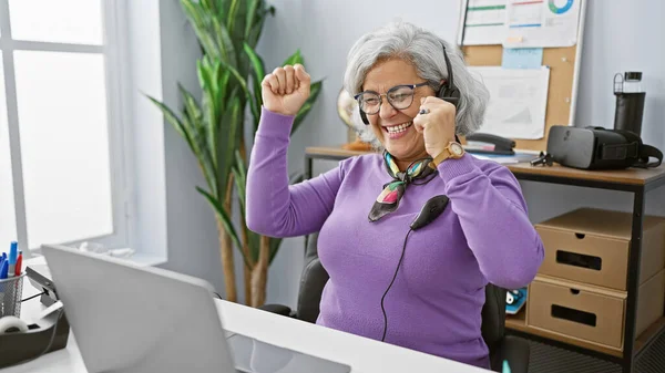 陽気な成熟した女性は 現代のオフィスのセットアップで働いている間 ヘッドセットを着用して成功を祝います — ストック写真