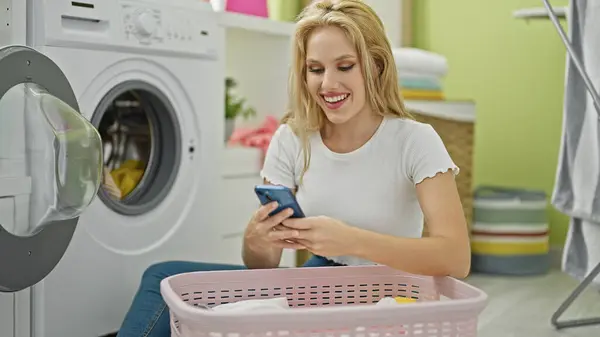 洗濯室でスマートフォンを微笑んで使用する若い金髪の女性 — ストック写真