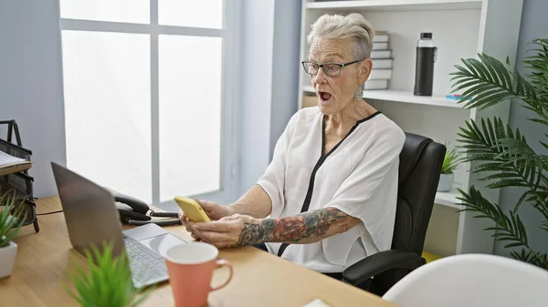 Wow! grey-haired senior woman business worker shocked at her office while using smartphone